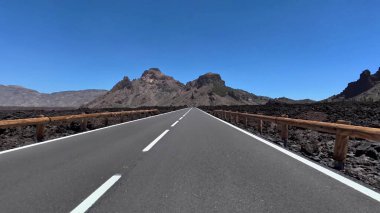 Büyüleyici ve manzaralı bir yol berrak mavi gökyüzünün altında uzanan çarpıcı volkanik bir arazide uzanır.