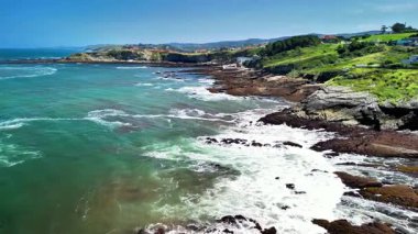 Dalgaların kayalık kıyılara çarptığı, yemyeşil tepelerin yemyeşil olduğu çarpıcı bir kıyı manzarası.