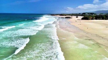 Sakin bir sahili gösteren nefes kesici bir hava çekimi. Dalgaların dalgalı bir gökyüzünün altında yumuşak kumu öptüğü yer.