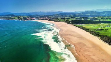 Hava manzarasında çarpıcı bir sahil, çarpan dalgalar ve yemyeşil tepeler var.