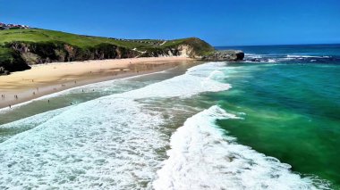Güzel bir plaj sahnesinde kuma vuran dalgalar ve yemyeşil tepeler arka planda yükseliyor.