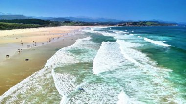 Göz kamaştırıcı ve pitoresk bir plaj sahnesi yumuşak dalgalar, yumuşak altın kum ve açık mavi gökyüzünü içerir.