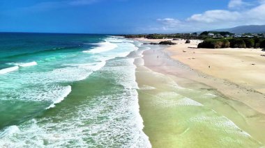 Sakin bir sahili gösteren nefes kesici bir hava çekimi. Dalgaların dalgalı bir gökyüzünün altında yumuşak kumu öptüğü yer.