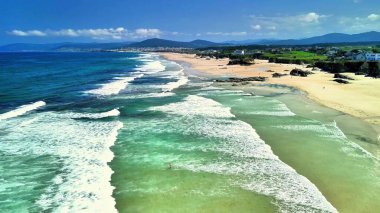 Nefes kesici bir hava manzarası berrak mavi gökyüzünün altında sakin sahili ve canlı denizi güzel bir şekilde gösteriyor.