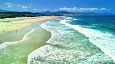 Nefes kesen hava perspektifi açık mavi gökyüzünün altında kumlu bir sahile çarpan dalgaları gösteriyor.