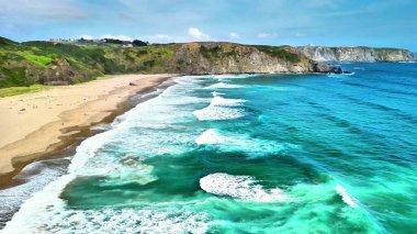 Temiz turkuaz suları, el değmemiş beyaz kumları ve arka planda yemyeşil kayalıkları olan nefes kesici bir sahil manzarası.