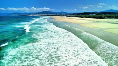 Berrak turkuaz sular, yumuşak dalgalar ve parlak güneşli bir gökyüzünün yer aldığı güzel ve huzurlu bir sahil manzarası.