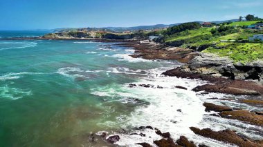 Dalgaların kayalık kıyılara çarptığı, yemyeşil tepelerin yemyeşil olduğu çarpıcı bir kıyı manzarası.