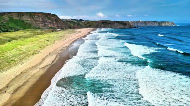 Çarpıcı bir hava manzarası sakin okyanus dalgalarını serin bir plajda gösteriyor, yemyeşil ve güzelliklerle çevrili.