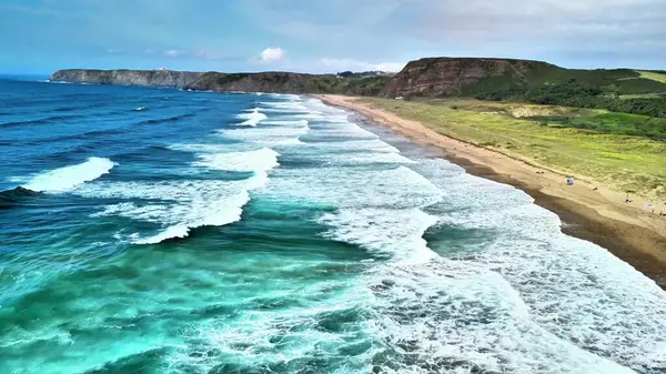 Nefes kesici hava sahneleri sakin okyanus dalgalarının görkemli kayalıklarla çevrili kumlu bir plaja çarptığını gösteriyor.