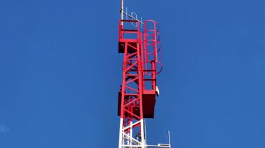 Geniş ve detaylı bir manzara, açık mavi gökyüzüne karşı uzun kırmızı ve beyaz bir telekomünikasyon kulesini gösteriyor.