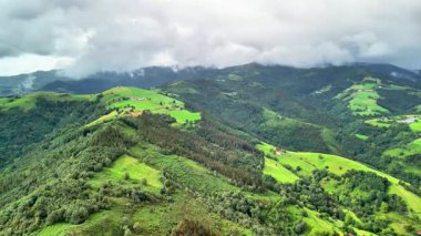 Göz kamaştırıcı bir hava manzarası yemyeşil dağları ve yuvarlanan tepeleri sergiliyor bulutlu bir gökyüzünün altında.