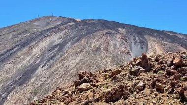 Göz kamaştırıcı manzara, berrak mavi gökyüzünün altında görkemli bir volkanik dağ ve kayalık araziyi içerir.