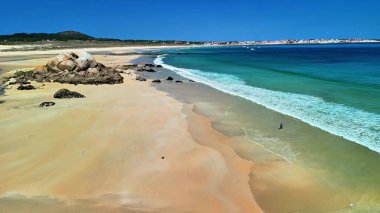 Güzel sahil manzarası turkuaz dalgaların açık mavi gökyüzü altında kayalık oluşumlara çarptığını gösteriyor.