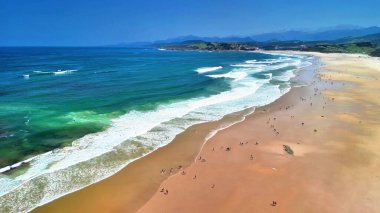 Güneşli bir plajın güzelliğini tecrübe edin, dalgalar çarpıyor, sörfçüler biniyor, ve insanlar sıcak kumun tadını çıkarıyor.