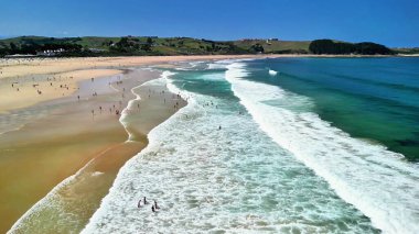 Göz kamaştırıcı bir hava manzarası güneşli bir plaj sahnesi yakalar, dalgaları gösterir ve insanlar okyanusun keyfini çıkarırlar.