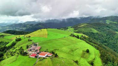Etkileyici çiftlikleri olan canlı yeşil tepelerin manzarası dramatik, girdap gibi bulutlu bir zemin üzerine kurulmuş.