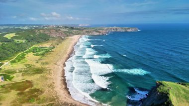 Sersemletici hava manzarası dalgalı okyanus dalgaları ve açık mavi gökyüzünün altındaki yemyeşil tepeleri gösteren güzel bir sahil.