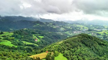 Sarsılan tepeleri ve çarpıcı bulutlu gökyüzünün altındaki canlı yemyeşil alanları gösteren nefes kesici bir hava manzarası.
