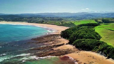Hava manzarası, yumuşak kumları, yeşil tepeleri ve rahatlamak için mükemmel mavi suları olan sakin bir sahili gösteriyor.