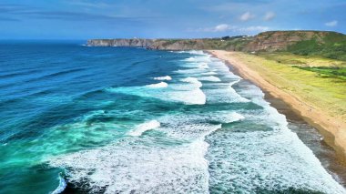 Çevresindeki yemyeşil kayalıklarla birlikte canlı ve yuvarlanan dalgaları gösteren nefes kesici bir deniz manzarası.