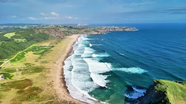 Sersemletici hava manzarası dalgalı okyanus dalgaları ve açık mavi gökyüzünün altındaki yemyeşil tepeleri gösteren güzel bir sahil.