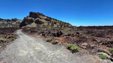 Berrak mavi gökyüzünün altındaki uzak bir dağla çarpıcı volkanik bir arazide manzaralı bir toprak yol esiyor.
