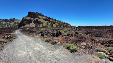 Berrak mavi gökyüzünün altındaki uzak bir dağla çarpıcı volkanik bir arazide manzaralı bir toprak yol esiyor.