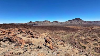 Geniş ve nefes kesici çöl arazisi, açık güneşli bir gökyüzünün altında kayalık oluşumlar ve görkemli dağları barındırır.