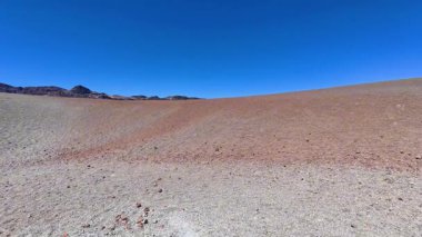 Göz kamaştırıcı bir manzara, açık mavi gökyüzünün altında canlı kırmızı ve gri toprak ile kuru ve çorak bir manzara sergiliyor.