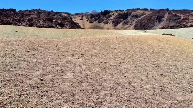 Kuru zemin ve açık mavi gökyüzünün altındaki kayalık tepelerle nefes kesici kurak bir manzara.