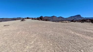 Berrak mavi gökyüzünün altındaki görkemli dağlarla kuru ve kayalık bir arazinin çarpıcı panoramik manzarası.