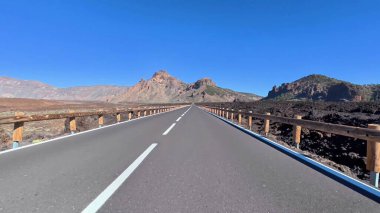 Büyüleyici bir yol, yukarıdaki açık mavi gökyüzünün altında çarpıcı volkanik bir arazide güzelce uzanır.