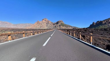 Büyüleyici bir yol sahnesi geniş, berrak mavi gökyüzü altında nefes kesici volkanik oluşumlar sergiliyor.