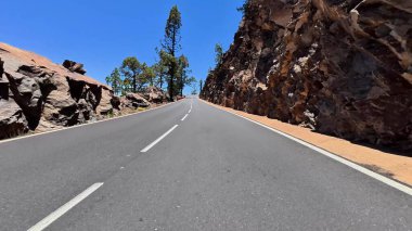 Manzaralı, pitoresk bir dağ yolu kayalık uçurumların ve açık mavi gökyüzünün altındaki yemyeşil ağaçların arasından esiyor.
