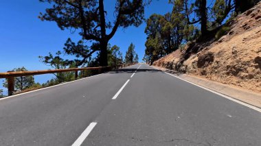 Sarmal bir dağ yolunun nefes kesici manzarası ağaçlarla kaplı, hepsi de yukarıdaki çarpıcı mavi gökyüzünün altında.