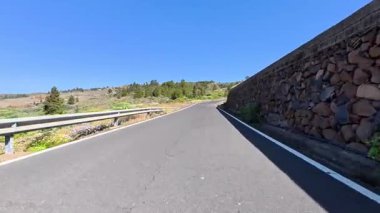 Etrafı canlı doğayla çevrili dolambaçlı bir dağ yolu parlak ve berrak bir mavi gökyüzünün altında uzanır.