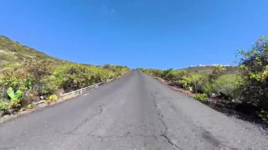 Parlak bir gökyüzüne karşı canlı bitki örtüsü tarafından çerçevelenmiş, yokuş yukarı uzanan sarmal bir yolun resimsel bir görüntüsü..