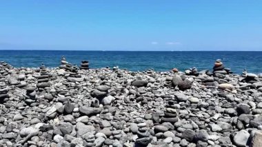 Altında sakin bir okyanus ve yukarıda parlak mavi bir gökyüzü olan engebeli bir sahilde istiflenmiş taşlardan huzurlu bir manzara.
