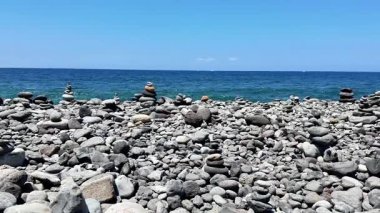 Güzel ve sakin okyanus manzarası açık mavi gökyüzünün altındaki kayalık sahilde sanatsal taş yığınları sergiliyor.