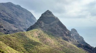 Bulutlu bir gökyüzünün altındaki canlı yeşil tepelerin arasında yer alan büyük bir dağın nefes kesici manzarası.