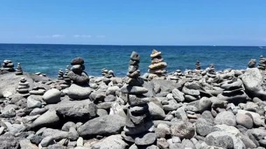 Sakin bir sahil sahnesinde kıyı şeridindeki dengeli taş yığınları parlak mavi gökyüzü ve tüylü bulutların altında yer alır.