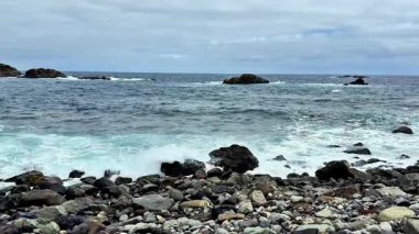 Bulutlu bir gökyüzünün altında engebeli kayalara çarpan dalgaların olduğu sakin ve sakin bir sahil manzarası.