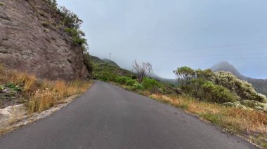 Geniş bulutlu gökyüzünün altında yeşil, engebeli kayalar ve uçurumlarla çevrili sakin bir dağ yolu.