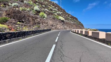 Güzel, dolambaçlı bir yol sahil boyunca ilerliyor, nefes kesici kaya oluşumları ve açık mavi gökyüzü sergiliyor.