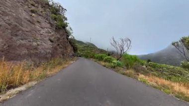 Geniş bulutlu gökyüzünün altında yeşil, engebeli kayalar ve uçurumlarla çevrili sakin bir dağ yolu.