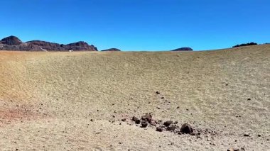 Kuru toprak ve dağınık kayalarla karakterize edilmiş engin kurak bir arazi parlak mavi gökyüzüne karşı kurulmuş.