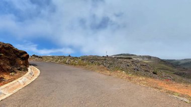 Güzel, dolambaçlı bir yol, tüylü bulutlar ve mavi gökyüzü ile çarpıcı bir dağ manzarası boyunca ilerliyor.