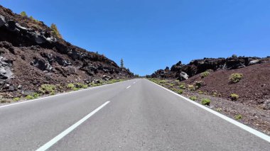 Berrak mavi gökyüzünün altında volkanik kayaların ve yemyeşil çimlerin arasından geçen bir otoyolun çarpıcı görüntüsü.