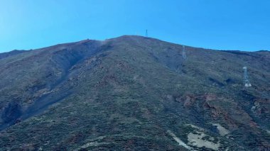 Açık mavi gökyüzünün altında haberleşme kulelerinin yer aldığı görkemli bir dağı gösteren nefes kesici bir manzara.